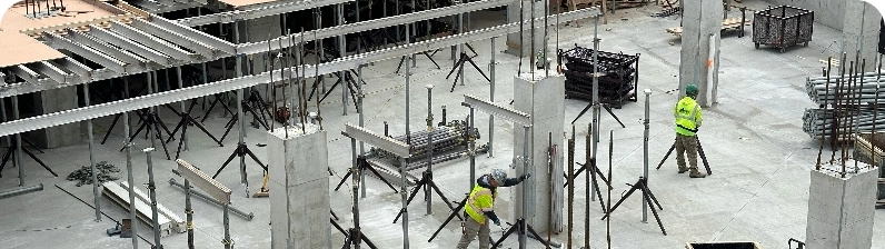 Construction site interior with concrete columns, scaffolding, and workers in high-visibility vests assembling steel formwork.