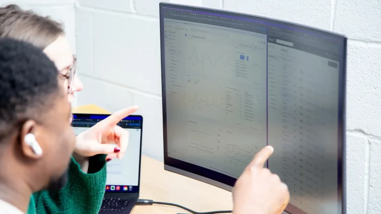 Two colleagues review data dashboards on a large monitor, pointing at charts and graphs in a bright office setting.