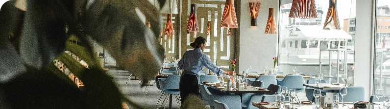 Server in a blue shirt sets a table in a modern waterfront restaurant with blue chairs and orange pendant lights, boats visible outside.