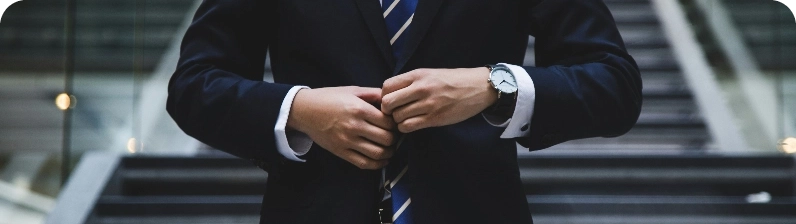 Close-up of a person buttoning a dark suit jacket, with a striped tie and a silver wristwatch visible on the wrist.